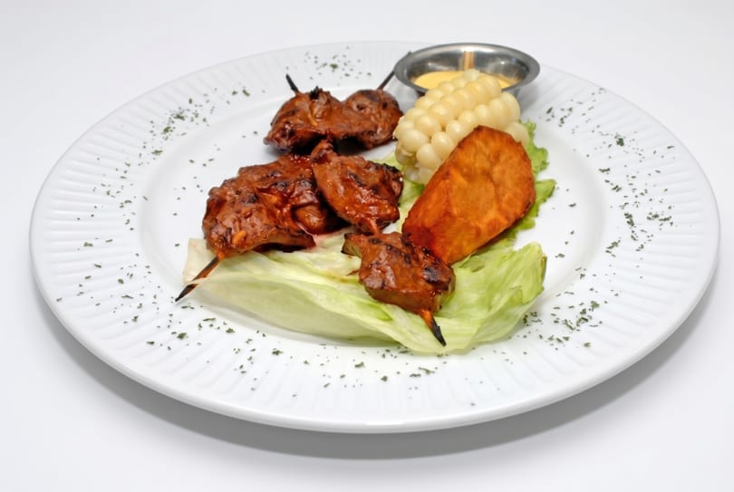 Anticuchos, a Peruvian dish, with grilled meat skewers, corn, sweet potato, and sauce on a white plate.